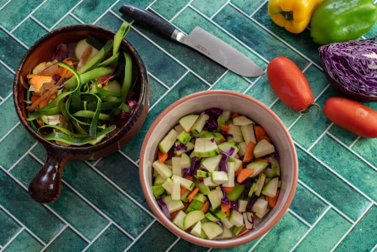 A close-up view of a ceramic bowl filled with freshly chopped vegetables — carrot, zucchini, and onion — next to a small pile of colorful vegetable scraps ready for compost. green tile background