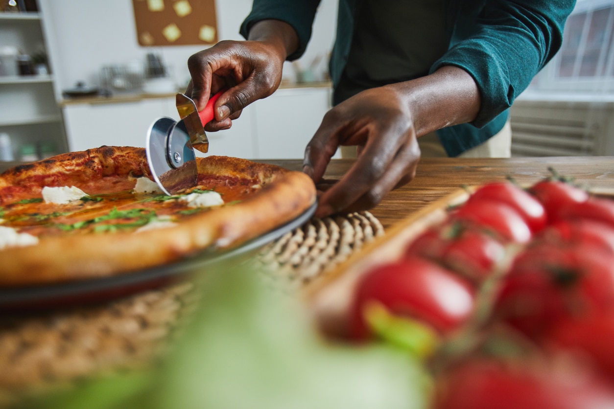 lose-up of African man standing at the table and cutting homemade pizza with knife in the kitchen